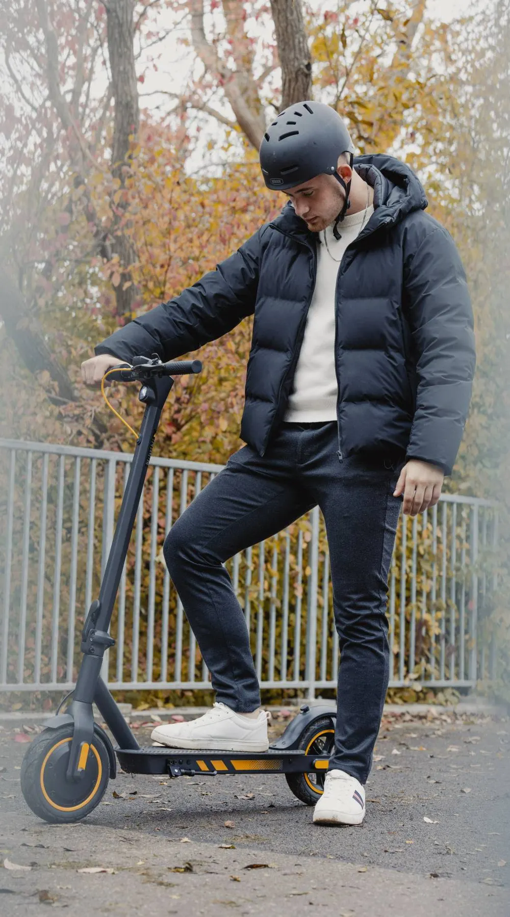Ung mand i sort dunjakke og hjelm sætter foden på ståbrættet af et sort el-løbehjul en tåget efterårsdag ved et rækværk
