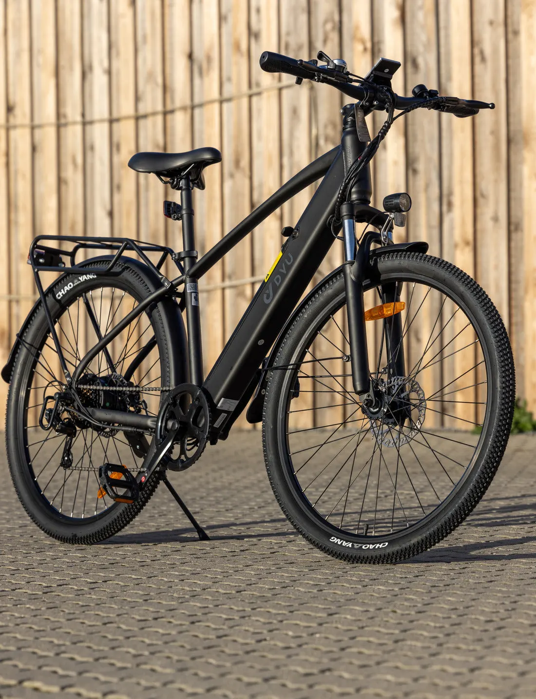 Sort DYU elcykel med batteriet integreret i rammen, bagagebærer, forgaffelaffjedring, skivebremser og forlygte, på støttebenet foran et træhegn i aftensol