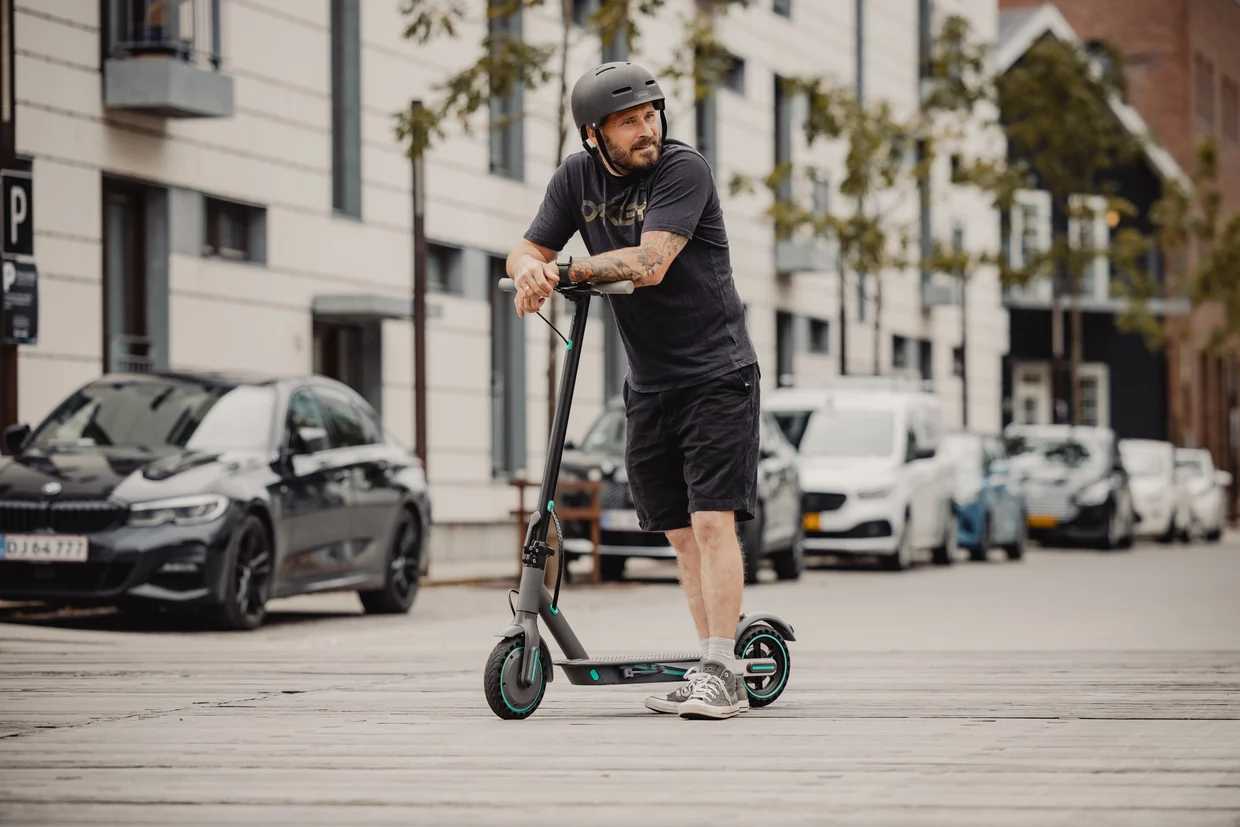 Mand med cykelhjelm læner sig op ad et sort el-løbehjul på en gade med parkerede biler