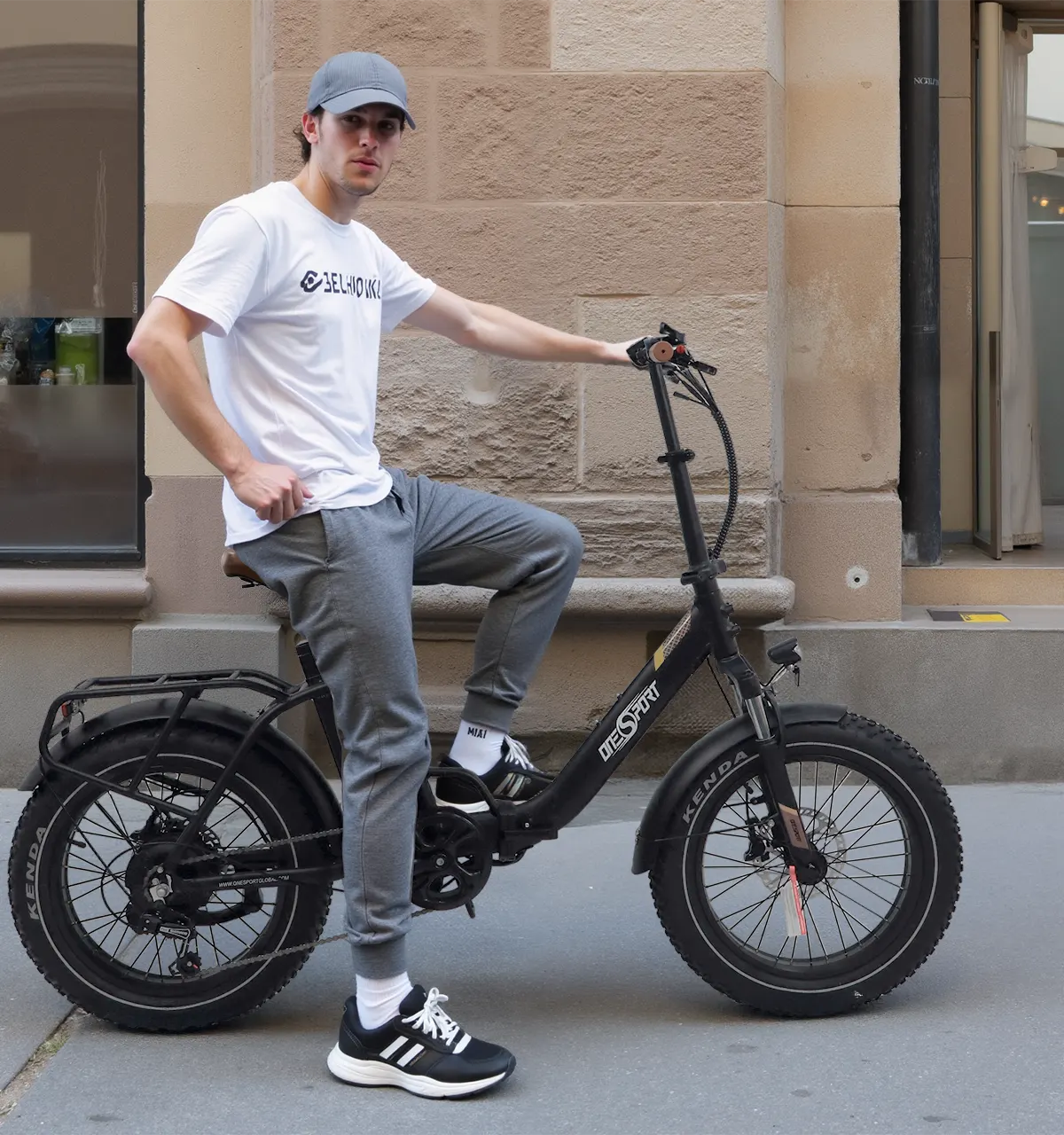 Ung mand med kasket står med en sort foldbar fat-tire elcykel med bagagebærer, forgaffelaffjedring og brede terrændæk på et fortov
