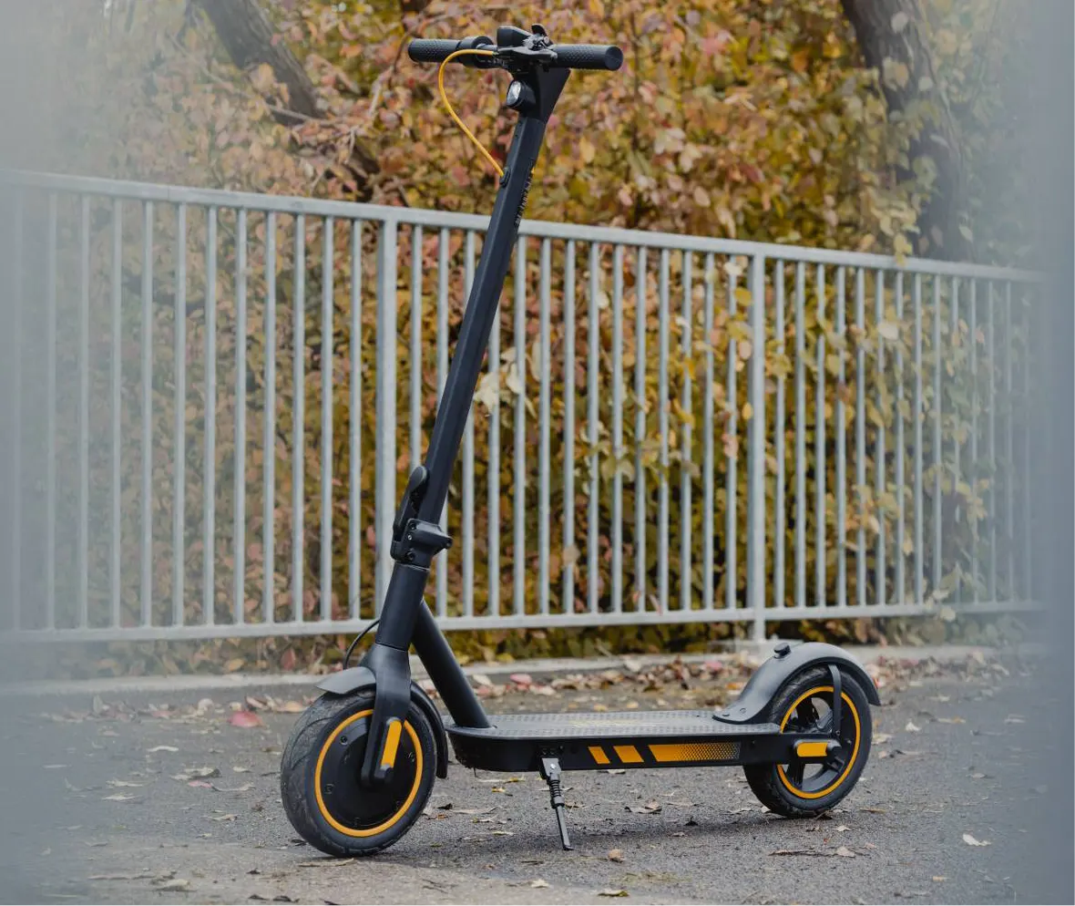 Sort el-løbehjul med orange fælge og synligt foldegreb midt på styrstammen, parkeret i tåget efterårsvejr ved et rækværk